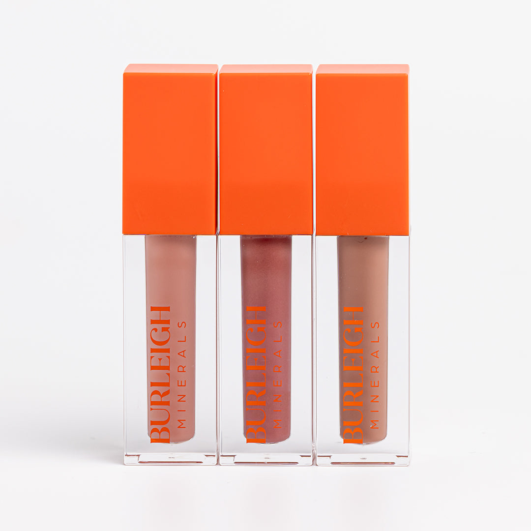 Three Liquid Lip Cremes lined up beside each other with an orange lid and clear container showing the product. On the left is shade Nude Peach, a pinky nude. In the middle is shade Rosey Mauve, a pinky mauve. And on the right is Almond Brown which is a deeper, neutral brown.