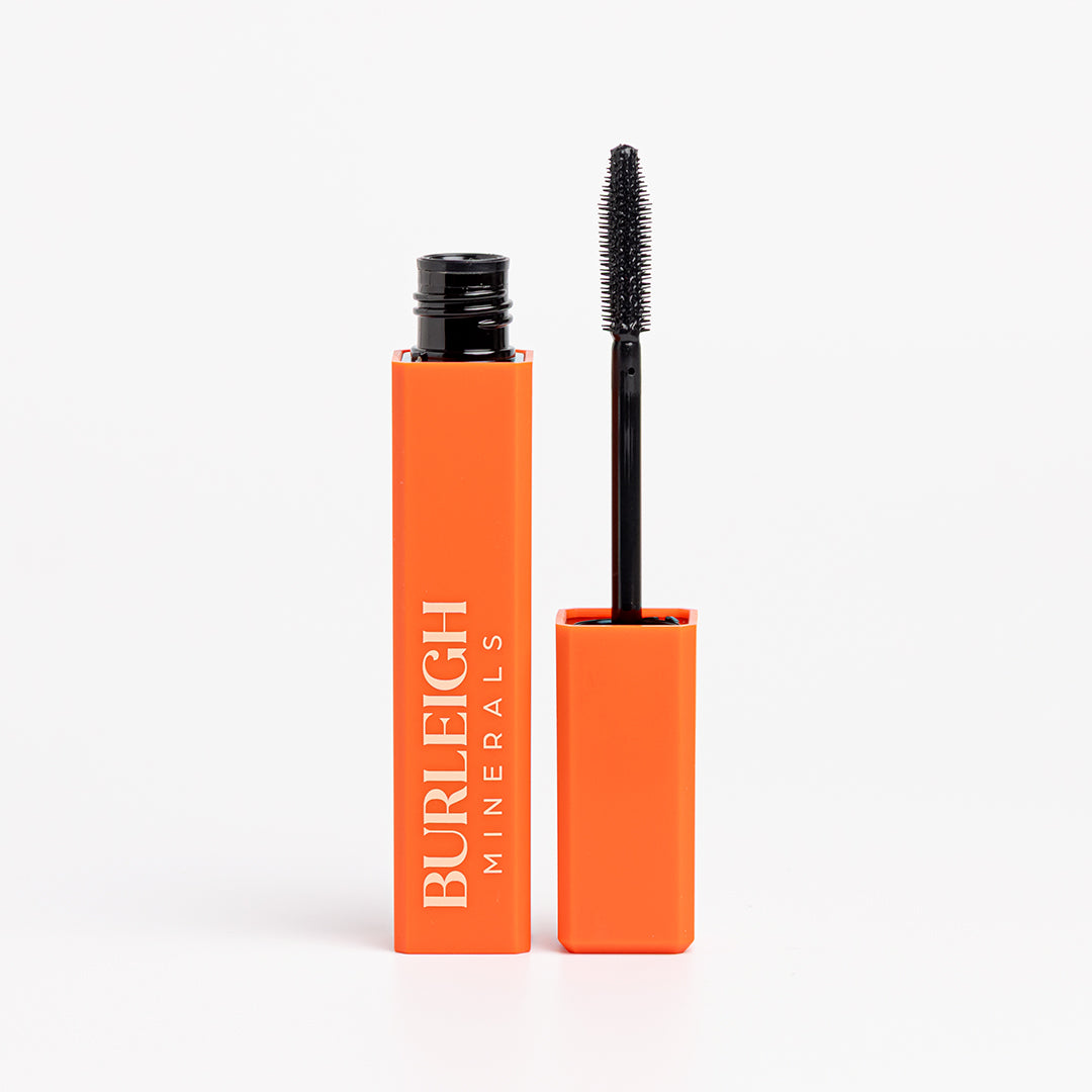 Water-resistant, black mineral mascara in bright orange packaging with the lid off and mascara wand pointing upwards, photographed against a white background.